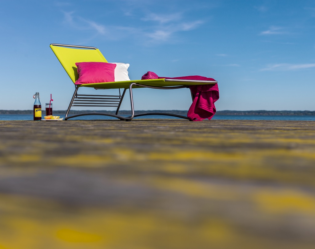 Grüne Weishäupl Sonnenliege mit roten Akzenten, Kissen und Getränk am Seeufer.