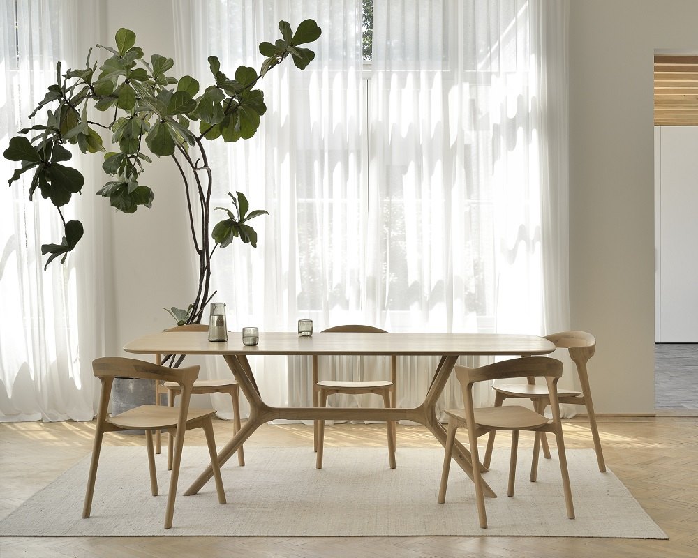 Ethnicraft X Dining Table Esstisch aus Eiche mit Stühlen und Pflanze vor hellem Fenster.