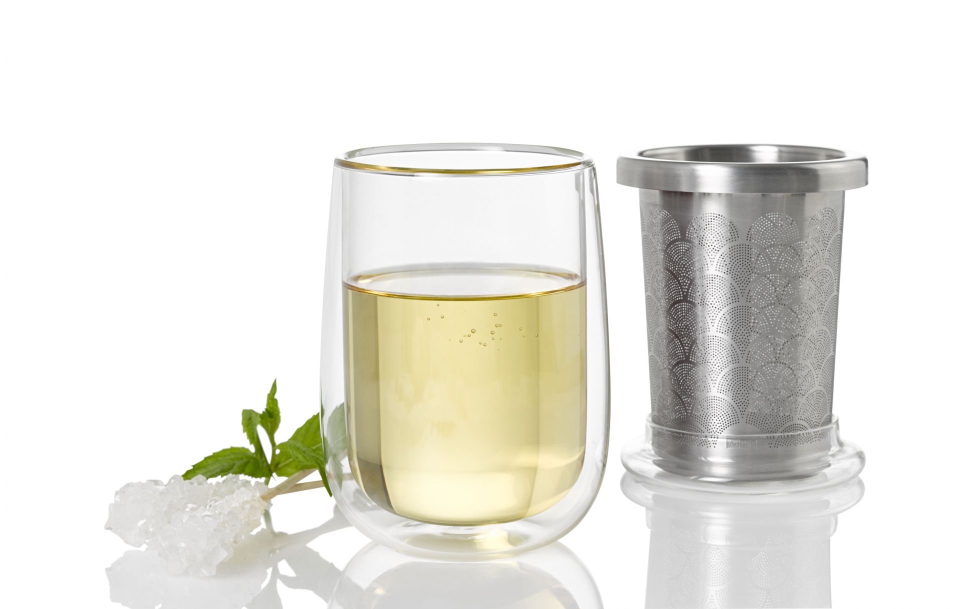 Teeglas mit frischem Tee und Teefilter aus Edelstahl, dazu Kandiszucker und Minze.