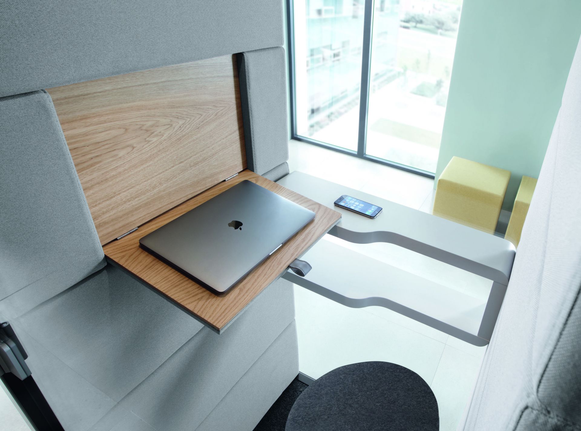 Laptop auf ausklappbarem Holzschreibtisch in grauer, schallisolierter Bürozelle mit Schreibtisch und Smartphone.